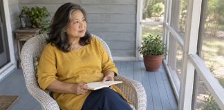 A woman in a mustard tunic holding an open notebook in a white wicker chair on a screened porch.