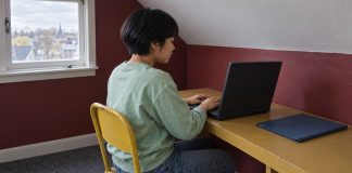 A woman in a mint sweatshirt typing on a black laptop at an ochre desk beside a closed blue folder.