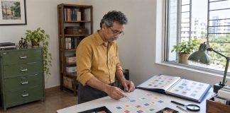A man in an ochre shirt using tweezers to arrange colorful stamps beside an open blue album at a work surface.