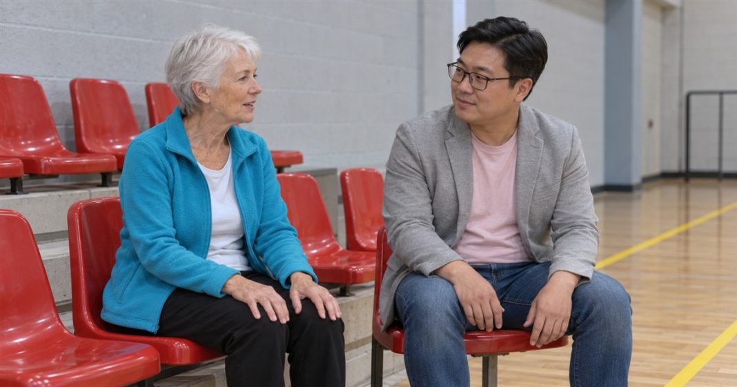 2726 realistic sports hall conversation 20261005 An older woman in a cyan cardigan and a man in a gray blazer talking on red seats in an indoor sports hall.