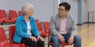 An older woman in a cyan cardigan and a man in a gray blazer talking on red seats in an indoor sports hall.