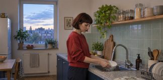 A woman in a red sweatshirt rinsing a white bowl at a kitchen sink with pale-green tiles.