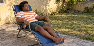 A man in an orange-and-white striped shirt resting in a blue folding recliner on a concrete backyard patio.