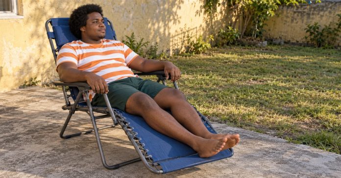 A man in an orange-and-white striped shirt resting in a blue folding recliner on a concrete backyard patio.