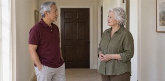 Two older adults standing apart and talking in a cream hallway near a closed dark-brown door.