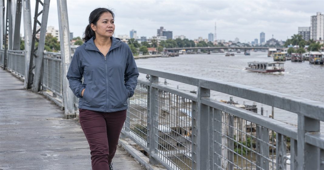 A woman in a blue-gray jacket and burgundy trousers walking alone on a steel pedestrian bridge beside an urban river.
