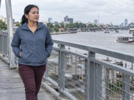 A woman in a blue-gray jacket and burgundy trousers walking alone on a steel pedestrian bridge beside an urban river.