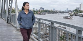 A woman in a blue-gray jacket and burgundy trousers walking alone on a steel pedestrian bridge beside an urban river.