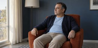 A man resting in a rust-orange armchair and looking toward daylight in a slate-blue sitting room.