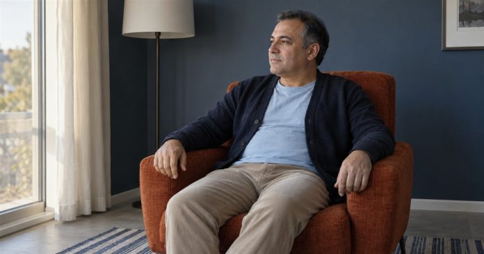 Present-day pause — ordinary rest A man resting in a rust-orange armchair and looking toward daylight in a slate-blue sitting room.