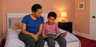 A caregiver and child looking at an open picture book together while seated on a bed in a warmly lit bedroom.