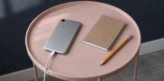 A silver phone connected to a white charging cable beside a closed notebook and yellow pencil on a pink table.