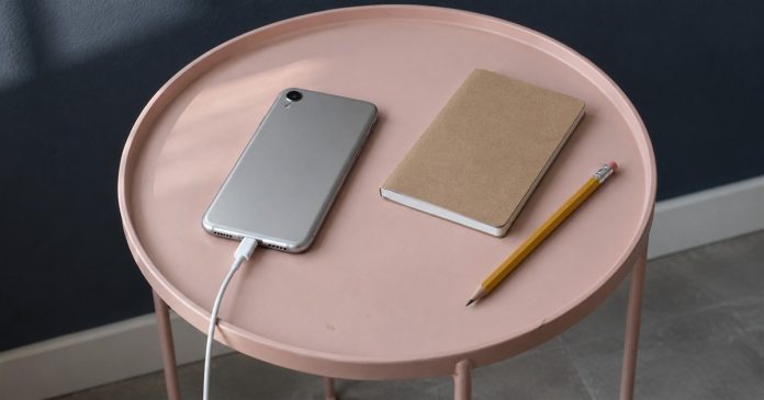 A silver phone connected to a white charging cable beside a closed notebook and yellow pencil on a pink table.