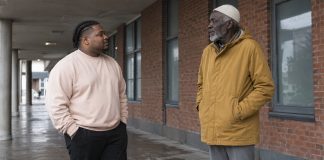 Two men in a pale sweatshirt and ochre coat talking beneath a covered brick walkway.
