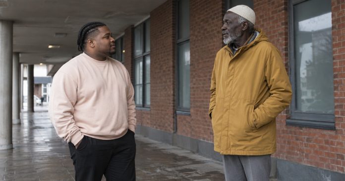 Changing friendships: brief public conversation — realistic cover 20261005 Two men in a pale sweatshirt and ochre coat talking beneath a covered brick walkway.