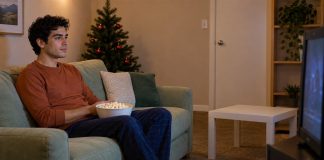 A man in an orange shirt watching television with popcorn on a green sofa beside a Christmas tree.