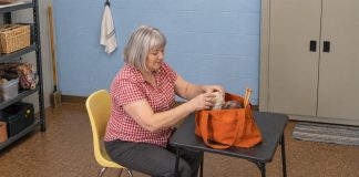 A silver-haired woman in a red gingham shirt arranging yarn in an orange tote on a folding table.