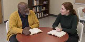 A man in a yellow cardigan and a woman in a green sweater discussing open books at a round table in a reading room.