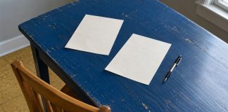 Two blank sheets of paper and a black-and-silver pen on a worn blue table beside a wooden chair.