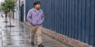 A man in a lavender jacket and khaki trousers walking along a damp sidewalk beside a blue corrugated wall.