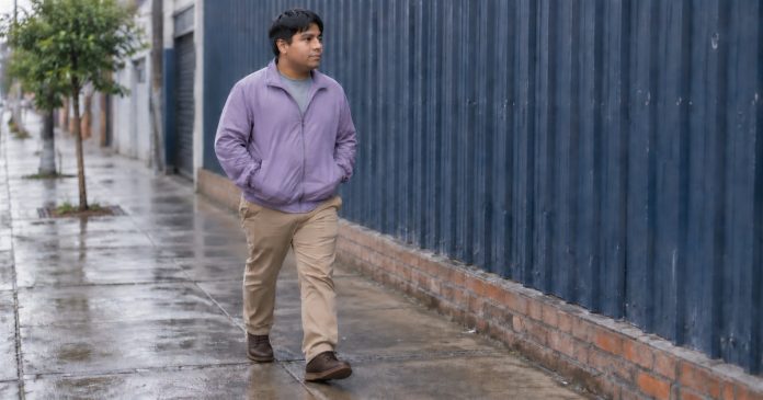 A man in a lavender jacket and khaki trousers walking along a damp sidewalk beside a blue corrugated wall.