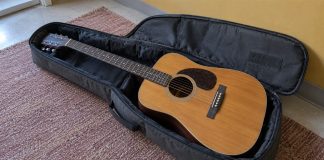 An acoustic guitar rests in an open dark gray soft case on a dusty rose rug beside a mustard-yellow wall.