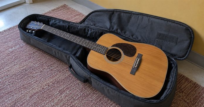 Getting back on track — Guitar ready for practice An acoustic guitar rests in an open dark gray soft case on a dusty rose rug beside a mustard-yellow wall.