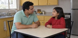 A man in a mint-green polo listening to a child in a red sweatshirt at a kitchen table.