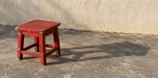 A worn red wooden stool casts a long shadow across a concrete floor beside a cream stucco wall.