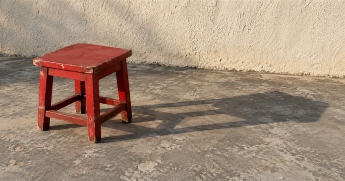Acknowledging Your Shadow — Red stool and ordinary shadow A worn red wooden stool casts a long shadow across a concrete floor beside a cream stucco wall.