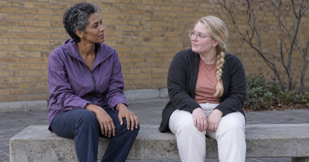 Quiet courtyard conversation — Respect and support Two women seated apart on a concrete courtyard bench, looking toward each other with their hands resting on their laps.