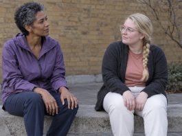 Two women seated apart on a concrete courtyard bench, looking toward each other with their hands resting on their laps.