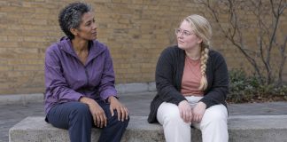 Two women seated apart on a concrete courtyard bench, looking toward each other with their hands resting on their laps.