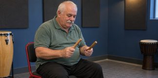 An older man seated on a red-framed chair, holding two wooden percussion sticks apart in a blue music-practice room.