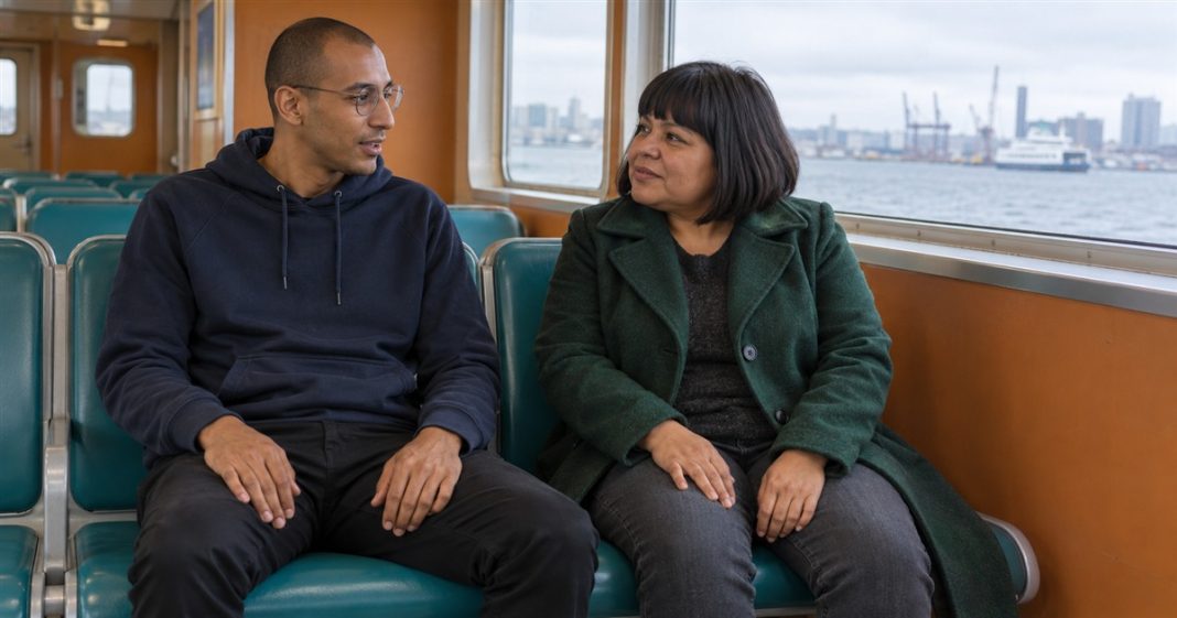 True Love Myths — Everyday Ferry Conversation Photographic Cover Man in a navy hoodie and woman in a green coat talking on teal ferry seats beside a harbor window.