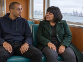 Man in a navy hoodie and woman in a green coat talking on teal ferry seats beside a harbor window.