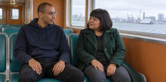 Man in a navy hoodie and woman in a green coat talking on teal ferry seats beside a harbor window.