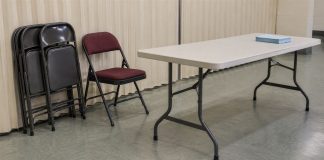 Folded metal chairs beside a burgundy chair and an off-white folding table with blue sheets in a plain community room.