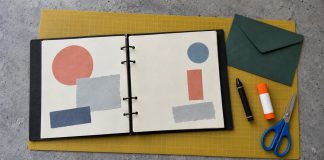 Open ring-bound scrapbook with colored paper shapes, green envelope, black crayon, glue stick and blue scissors on a yellow craft mat.