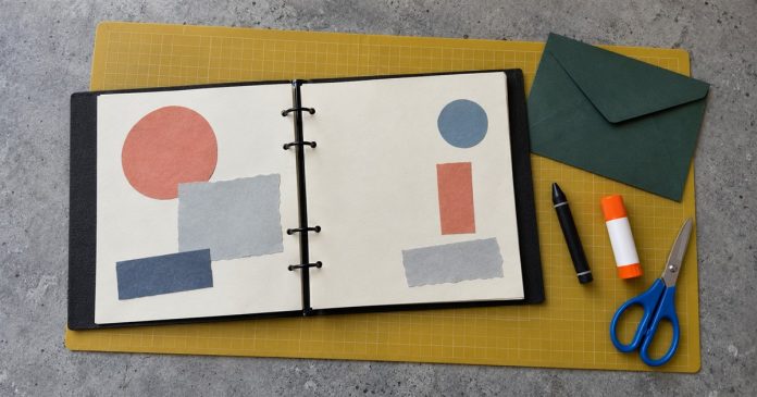 Clarity — Practical Scrapbook Workspace Photographic Cover Open ring-bound scrapbook with colored paper shapes, green envelope, black crayon, glue stick and blue scissors on a yellow craft mat.
