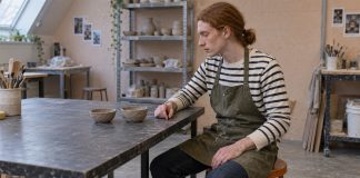 Red-haired man in a striped shirt and olive apron looking at two unfinished clay bowls on a workshop table.