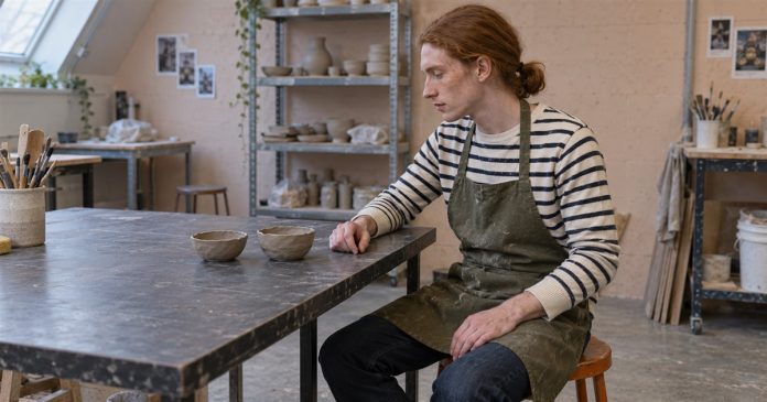 Listen to Intuition — Small Pottery Hobby Photographic Cover Red-haired man in a striped shirt and olive apron looking at two unfinished clay bowls on a workshop table.