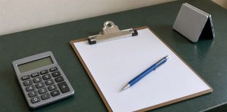 Photographic-style editorial image of a calculator, blank clipboard, blue pencil and folded silver device on a green desk.