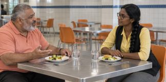 Photographic-style editorial image of two adults listening and talking over lunch at a cafeteria table.