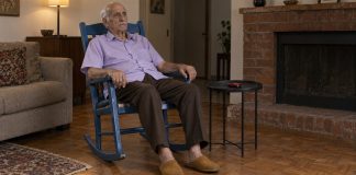 Photographic-style editorial image of an older man sitting in a blue rocking chair beside a closed red flip phone.