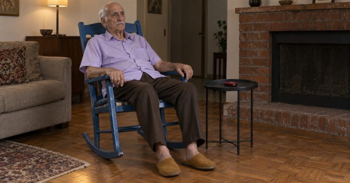 Photographic-style editorial image of an older man sitting in a blue rocking chair beside a closed red flip phone.