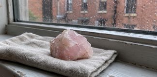 Photographic-style editorial image of rough pale-pink quartz on folded linen beside a rain-speckled window.