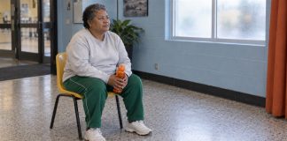 Photographic-style editorial image of a woman resting on a yellow chair with an orange water bottle in a quiet lobby.