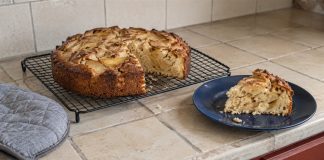 Homemade apple cake cooling on a wire rack, with a slice on a blue plate in a tiled kitchen.