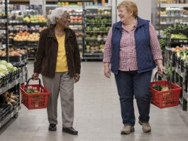 Two older friends smiling at each other while carrying grocery baskets through a supermarket produce aisle.